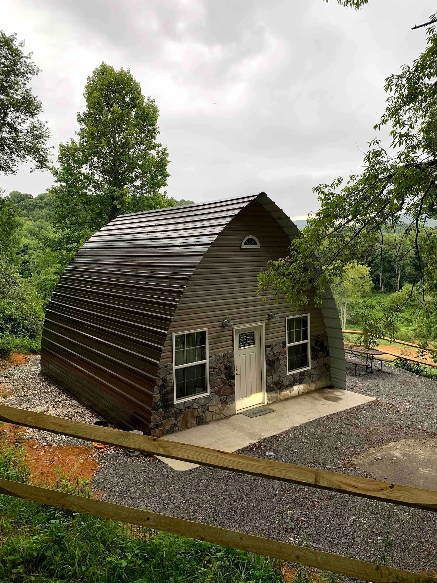 Dark pointed arch cabin with a masonry base and trees behind it.