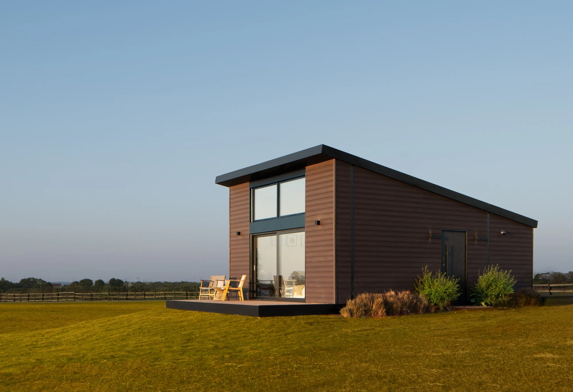 Published Unfold House dark-clad compact home with a glazed front facing an open grass field